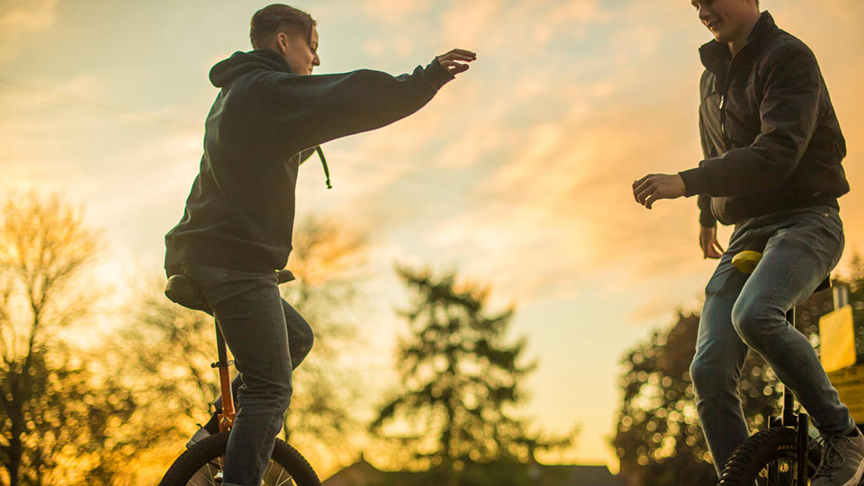 Two students on unicycles who are cycling near each other with a sun setting in the background